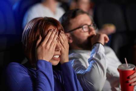 Selective focus of young scared girl covering eyes with hands, watching horror movie in cinema. Side view of scared bearded caucasian man and red headed woman enjoying frightening film.の写真素材
