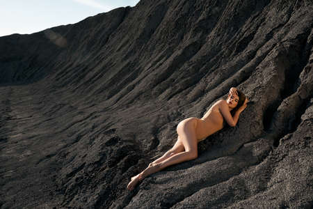 Side view of sensual caucasian female model wearing beige body posing in dry empty quarry in hot summer sunny day. Young stylish barefoot woman lying on black cracked ground and looking at camera.の写真素材
