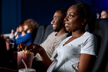 Selective focus of young woman watching film, eating popcorn with boyfriend in movie theater....