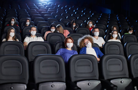 Front view of people enjoying film during world pandemic, looking at big screen. Young audience watching movie in cinema, wearing white face masks. Social distancing, pandemic concept.の写真素材