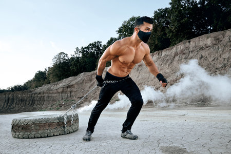 Strong active man with bare torso pulling big tyre on long chain outdoors. Muscular guy wearing protective face mask during pandemic. Concept of healthy lifestyle.の写真素材