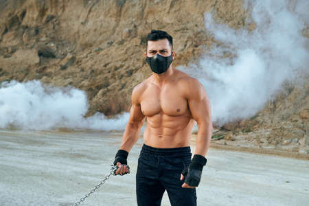 Front view of handsome man with athletic body holding metal chain in hands while standing at sand pit. Muscular male wearing sport pants, gloves and medical protective mask.の写真素材