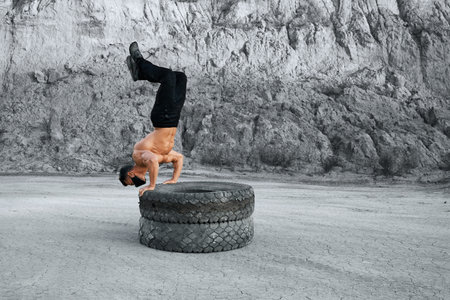 Strong man with bare torso doing push ups on two tyres at sand quarry. Active male wearing black...
