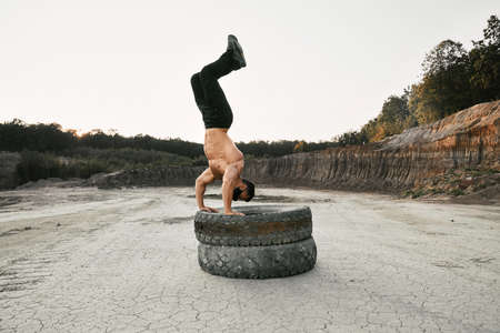 Healthy young man with athletic body using two heavy tyres for sport activity at sand pit. Muscular shirtless man wearing black spot pants and protective face mask.の写真素材