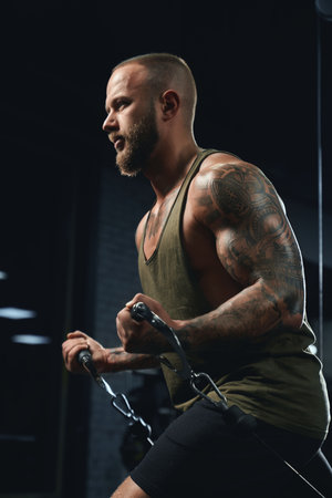 Portrait of muscular tattooed bodybuilder doing low cable crossover exercise. Side crop of man with...