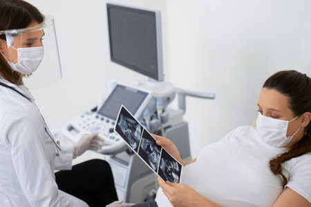 Happy pregnant woman in protective face mask looking at images of her unborn child after ultrasound screening. Female gynecologist sitting near and also wearing medical maskの写真素材