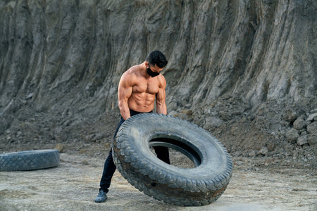 Professional bodybuilder in protective mask doing exercises with large black wheel at sand pit. Young man with naked chest leading healthy and active lifestyle.の写真素材
