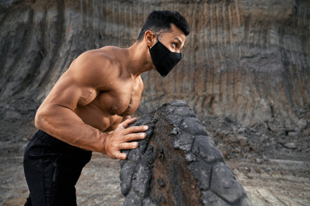 Side view of strong man in black face mask using heavy tyre for training on fresh air. Young guy with athletic body workout alone outdoors during pandemic time.の写真素材