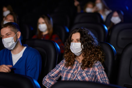 Side view of man and woman with curly brown hair enjoying date during world pandemic. Selective focus of young couple watching movie in cinema, wearing white face masks, social distancing.の写真素材