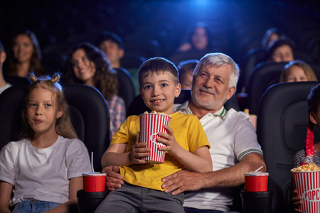 Selective focus of bearded senior grandfather enjoying time with grandson, kid sitting on knees and holding popcorn bucket. Happy caucasian family sitting in cinema, watching funny cartoon.の写真素材