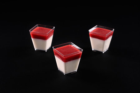 Front view of three small square glasses with milky panna cotta and strawberry jam. Sweet delicious dessert decorated with bright red topping, isolated on black background. Food concept.の写真素材