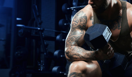 Incognito male bodybuilder training biceps with dumbbell. Close up of sportsman with perfect tensed muscular body posing in gym in dark atmosphere, sitting on bench. Concept of bodybuilding.の写真素材
