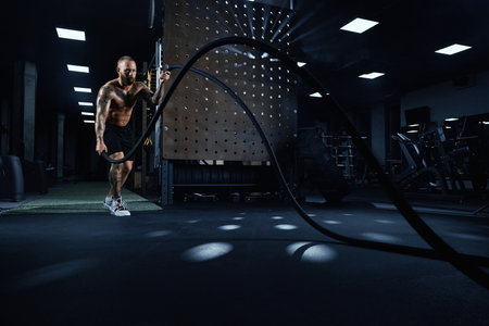 Front view of muscular brunette shirtless man doing battle rope training in gym in dark atmosphere....
