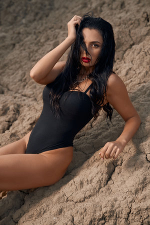 Adorable young lady dressed in only black bodysuit lying on dry sand among empty quarry. Fashion model having bright makeup and dark wavy hair.の写真素材