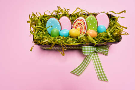 Colorful easter eggs in wooden basket with green bow and gingerbread cookies isolated on pink background. Holiday decoration, gift box.の写真素材