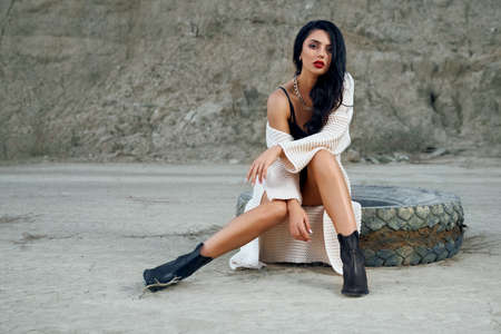 Stunning young lady with long dark hair dressed in trendy bodywear posing among empty sand quarry. Fashion model sitting old big tyre outdoors and looking at camera.の写真素材