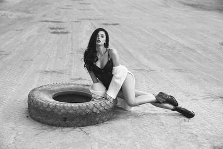 Black and white image of sexy young lady in stylish black bodywear posing near old car tyre outdoors. Fashion model with bright makeup and long dark hair sitting among empty quarry.の写真素材
