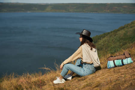 Attractive female traveler with backpack sitting on high hill and looking at Dniester river. Concept of active lifestyle and nature beauty.の写真素材