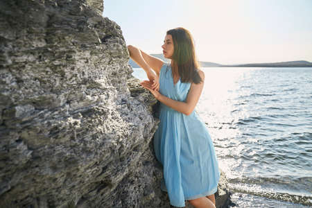 Pleasant young woman with brown hair leaning on rock near Dniester river. Fashion model in stylish dress posing outdoors during sunny weather.の写真素材