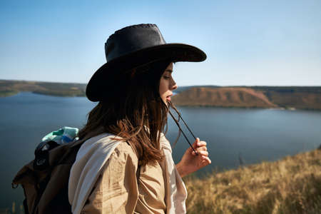 Side view of pretty woman in cowboy hat with backpack spending leisure time on fresh air. Attractive traveler hiking alone at national park Podillya Tovtry.の写真素材