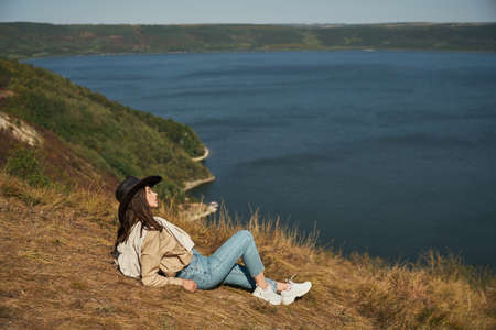 Relaxed woman in casual outfit and cowboy hat sitting on green hill and looking at Bakota bay. Young...