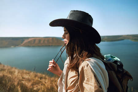 Satisfied young woman in casual outfit and cowboy hat posing on high green hill at Bakota area. Amazing view of Dniester river background.の写真素材
