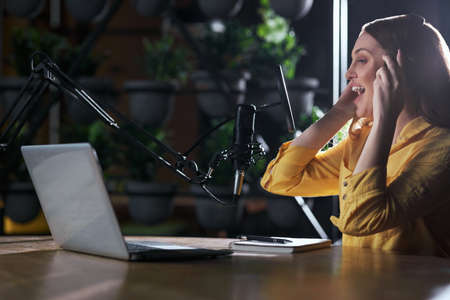 Side view of young beautiful woman in stylish yellow shirt singing into modern microphone near...