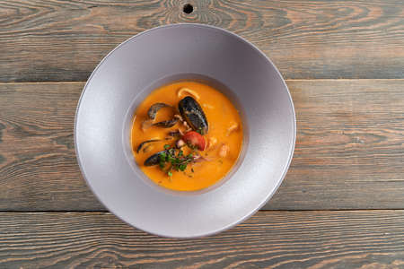 From above view of tasty bright orange soup with mussels in black shells in white plate on wooden restaurant table. Delicious seafood served with sprouts and tomatoes. Concept of exotic asian cuisine.の写真素材