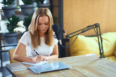 Front view of young attractive woman sitting at the table and writing some information in notebook....