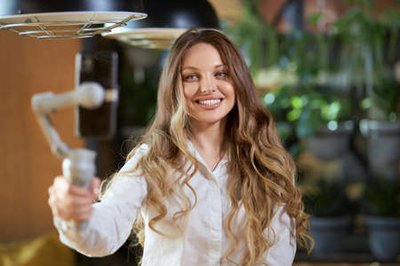Close up portrait of cute and beautiful smiling woman in white shirt holding selfie stick and...