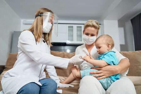 Female doctor in medical uniform and protective mask examining little baby that sitting on mother knees. Nurse checking cute patient at apartment.の写真素材