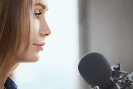 Close up of young attractive woman standing and recording some information in microphone online. Concept of process communication about different topics with modern microphone.の写真素材