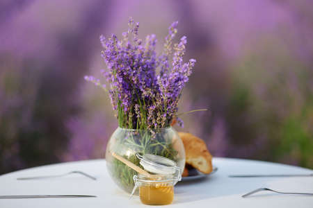 White wooden table decorated with fresh delicious croissants, honey jar with wooden spoon and glass vase with lavender bouquet. Beautiful decoration in lavender field full of blooming purple flowers.の写真素材