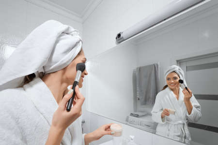 Side view of smiling attractive young woman doing makeup in bathroom looking at himself in the mirror. Concept of care body and preparing skin for makeup in hotel.の写真素材
