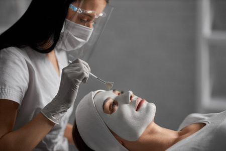 Side view of young beautiful woman lying on procedure for cleaning face in beautician in protective mask. Concept of special beauty procedure for improvements skin face and body.の写真素材