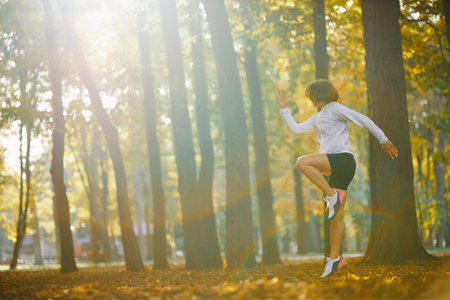 Side view of young woman in white sport jacket and black shorts jumping at park during morning workout. Concept of strength and sport activity.の写真素材