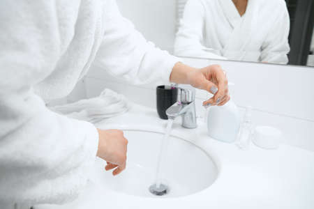 Side view of young woman in white robe standing near sink and washing hands with special...