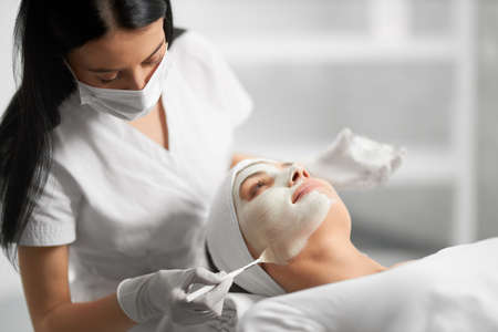 Side view of beautiful brunette beautician applying special white cream for face young cute patient. Concept of process procedure for improving face skin with cosmetics.の写真素材