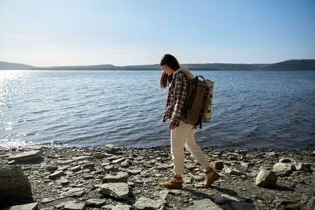 Full length portrait of beautiful young woman in casual wear with backpack walking along riverside....