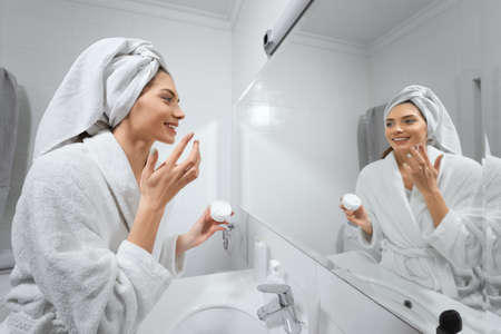 Side view of smiling cute young woman apply facial cream look in mirror. Concept of body care and preparing skin for makeup with good mood at home or hotel.の写真素材