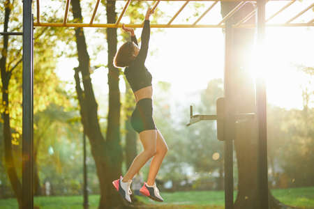 Young sports woman hanging on horizontal bar during workout on fresh air. Active brunette in sport...