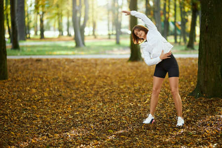 Beautiful young woman in activewear warming up at park before morning run. Healthy and fit brunette...