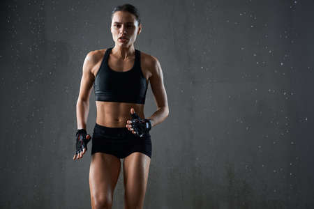 Portrait of wet female bodybuilder with perfect muscular body jogging on spot isolated in empty hall, loft interior. Concentrated athletic woman practicing hard cardio sport exercise under rain.の写真素材