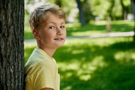 Cute caucasian boy in casual wear posing near tree at green park. Happy child spending free time on fresh air. Summer warm days.の写真素材