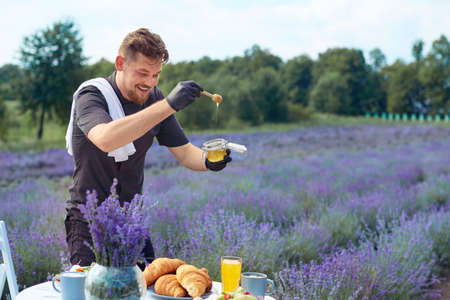 Side view of smiling caucasian waiter holding honey jar and wooden spoon outdoors. Young male chef...