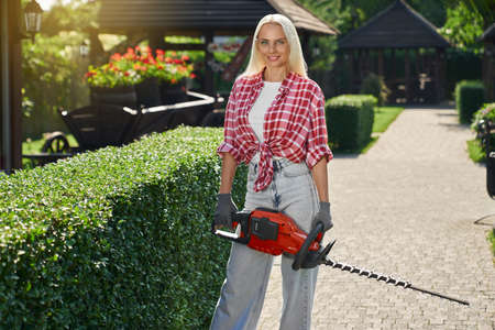Adorable female gardener in protective glasses and gloves posing on back yard with electric hedge trimmer and smiling on camera. Summer work at garden. Modern equipment.の写真素材