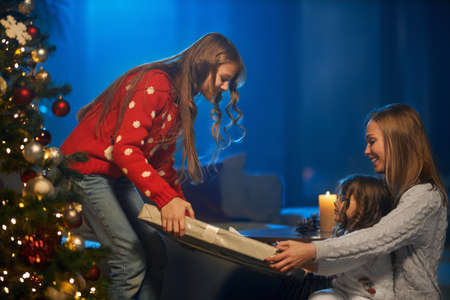 Happy caucasian mother and her little daughters sharing with presents while sitting on floor near christmas tree. Happy family celebrating winter holidays. Festive mood concept.の写真素材