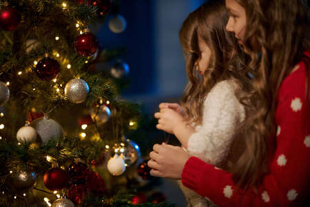 Side view of pretty caucasian little girls decorating christmas tree with colorful baubles and toys. Happy sisters preparing cozy home for christmas celebration.の写真素材