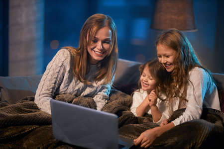 Two little sisters and their mother sitting on couch under soft blanket and using modern laptop for watching cartoons. Happy family spending evening time with pleasure.の写真素材
