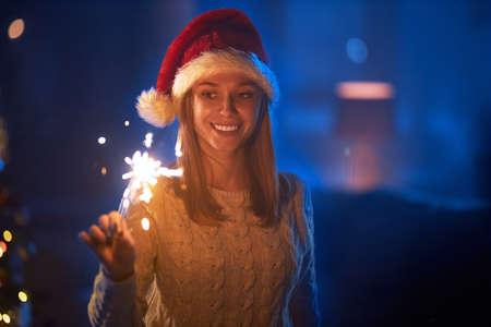 Happy caucasian woman in santa hat standing at dark living room with burning sparklers in hands. Pretty blonde celebrating new year of christmas at home. Concept of winter holidays.の写真素材